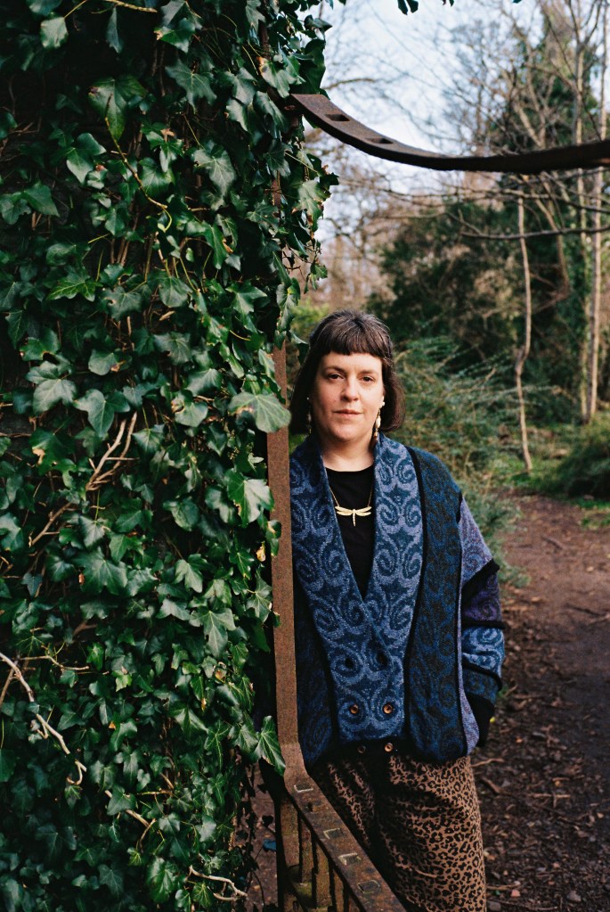 Photograph of a woman with blue cardigan and brown jeans standing by a rusty gate and an ivy hedge